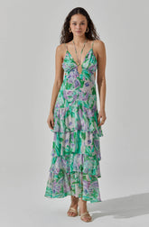 A woman is wearing a sleeveless, long summer dress with spaghetti straps. The dress features a vibrant floral pattern in shades of green, purple, and lavender, with a keyhole cutout at the front. It has a tiered, ruffled skirt that reaches down to her ankles.